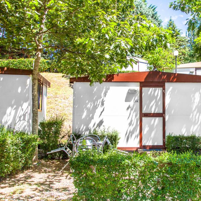 Small white cabin surrounded by trees and bushes with outdoor furniture.